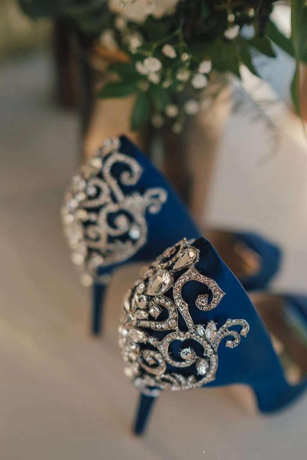 Close-up of navy blue high heels with ornate rhinestone embellishments on the back, with a blurred background of a flower arrangement in a silver vase.