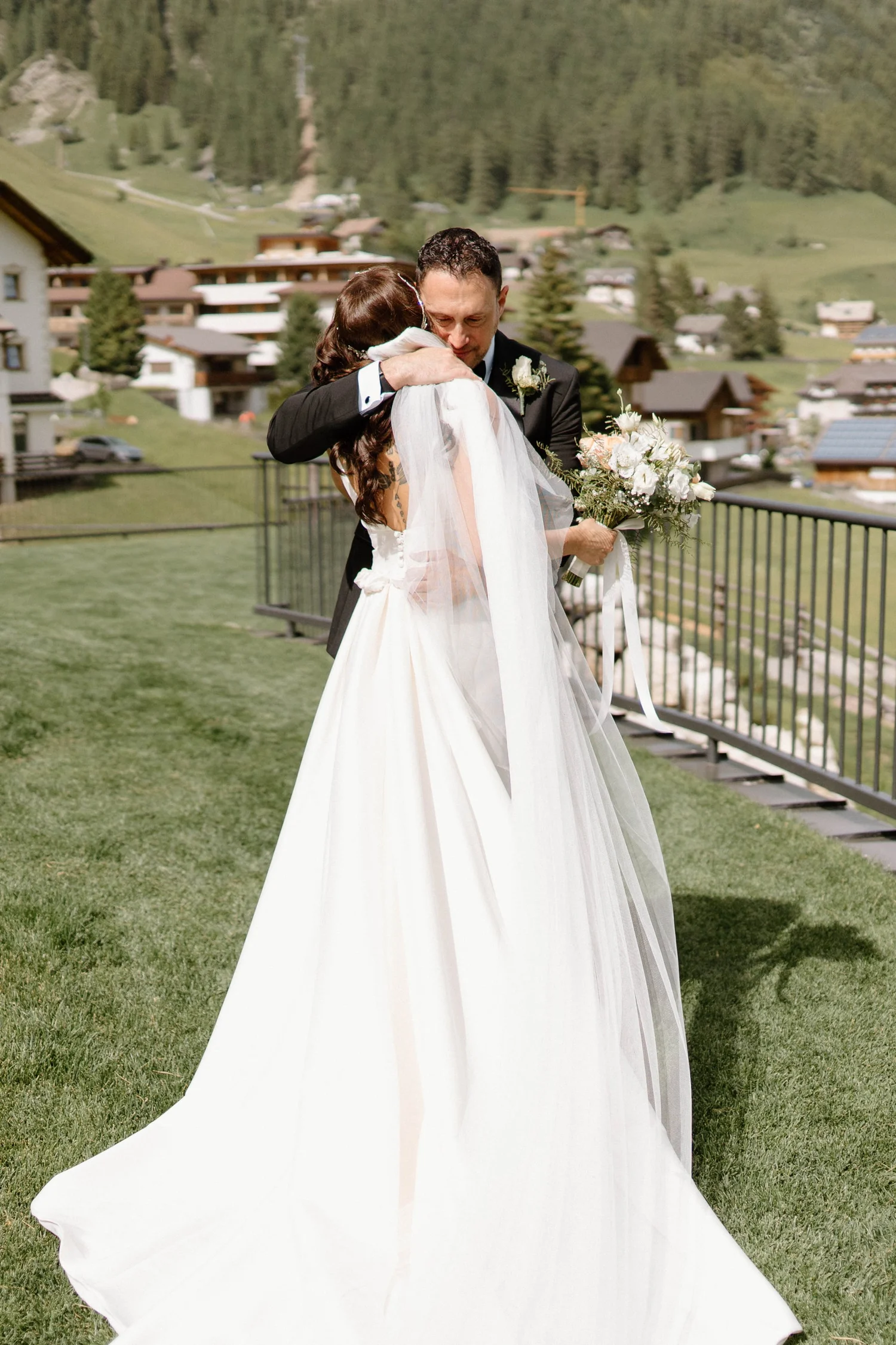 A bride and groom share a hug outdoors on a grassy area with a mountain village in the background. The bride is wearing a white wedding dress and veil, holding a bouquet of white flowers. The groom is dressed in a dark suit with a white shirt and bou