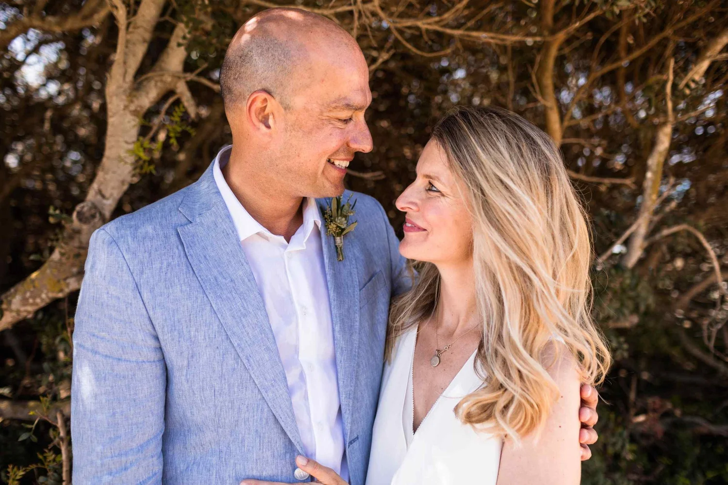A man and woman smiling and looking at each other, holding each other close outdoors in front of trees.