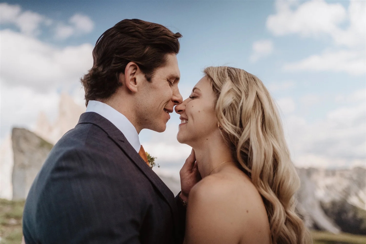 A close-up of a couple with their noses touching, smiling, outdoors on a cloudy day.