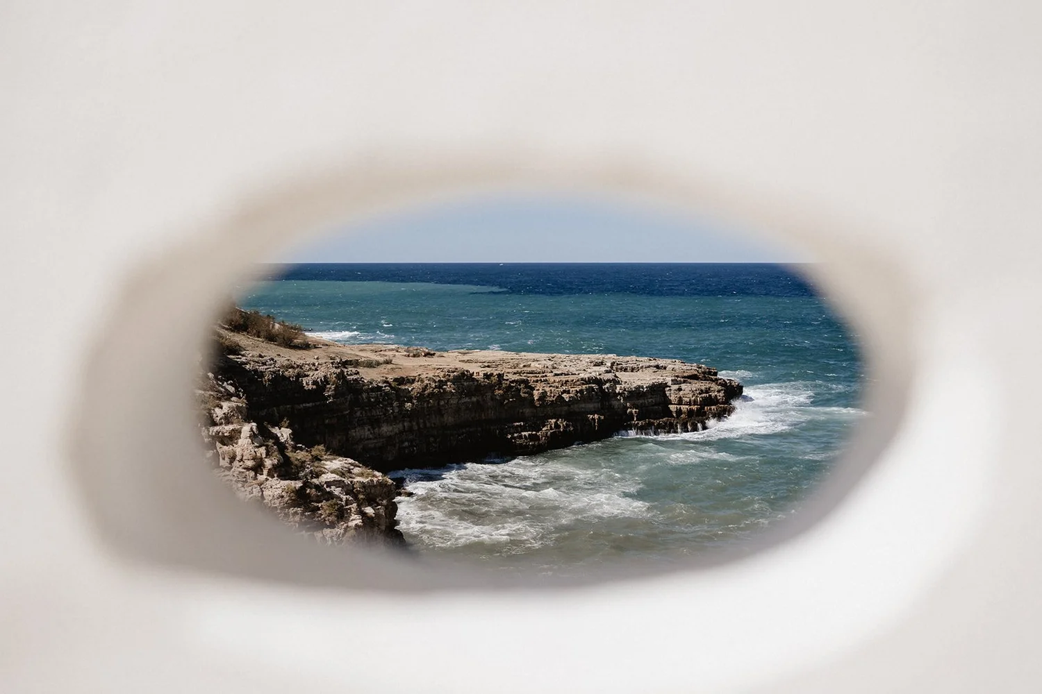 View of ocean and rocky coastline through a circular hole in white material.