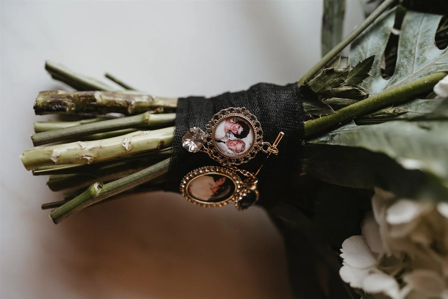 A bouquet of dried green and brown leaves and stems, with a black fabric ribbon tied around the stems. Attached to the ribbon is a pendant with a photo of two women and a child inside a decorative gold frame, and a large clear gemstone is also attach