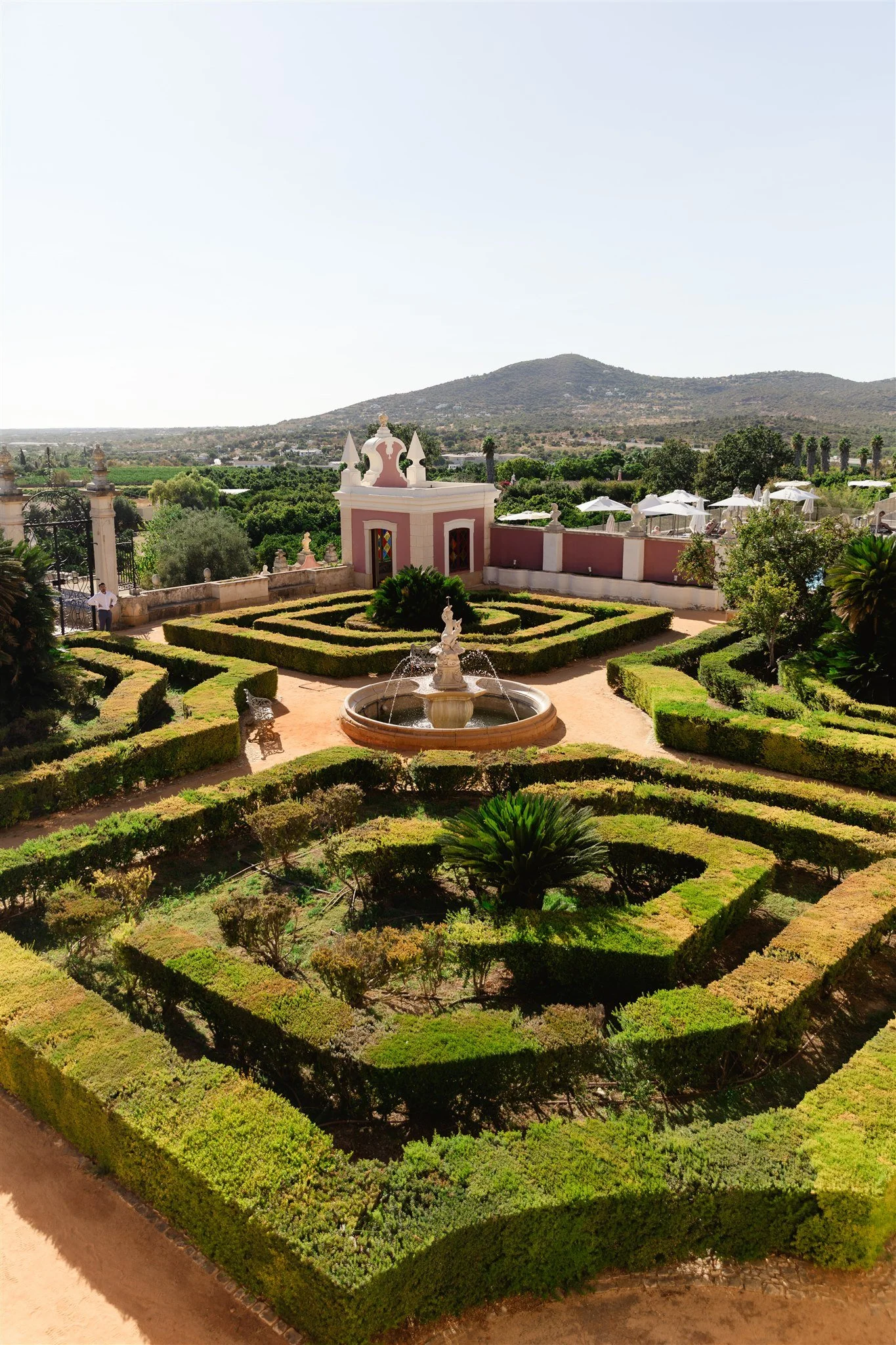 A formal garden with neatly trimmed hedges and pathways, a central fountain with statues, and a pink and white building with tower-like structures and stained glass windows. In the background, there are green trees, white umbrellas, and mountains und