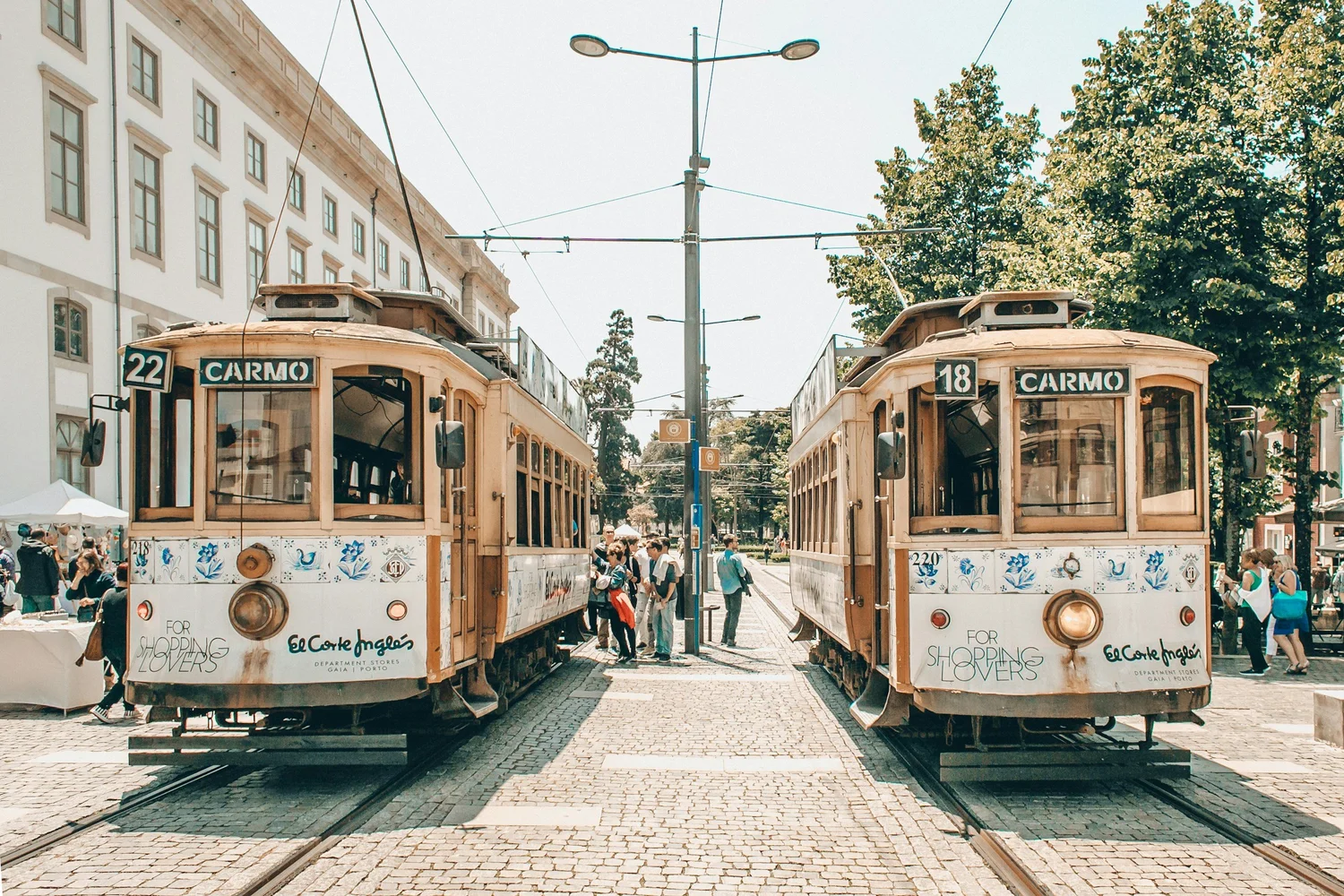 Two vintage trolley cars at a city intersection with people walking around, surrounded by buildings, trees, and street lights.