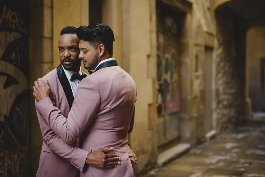 Two men in pink suits embracing each other in a narrow alley with brick walls and graffiti.