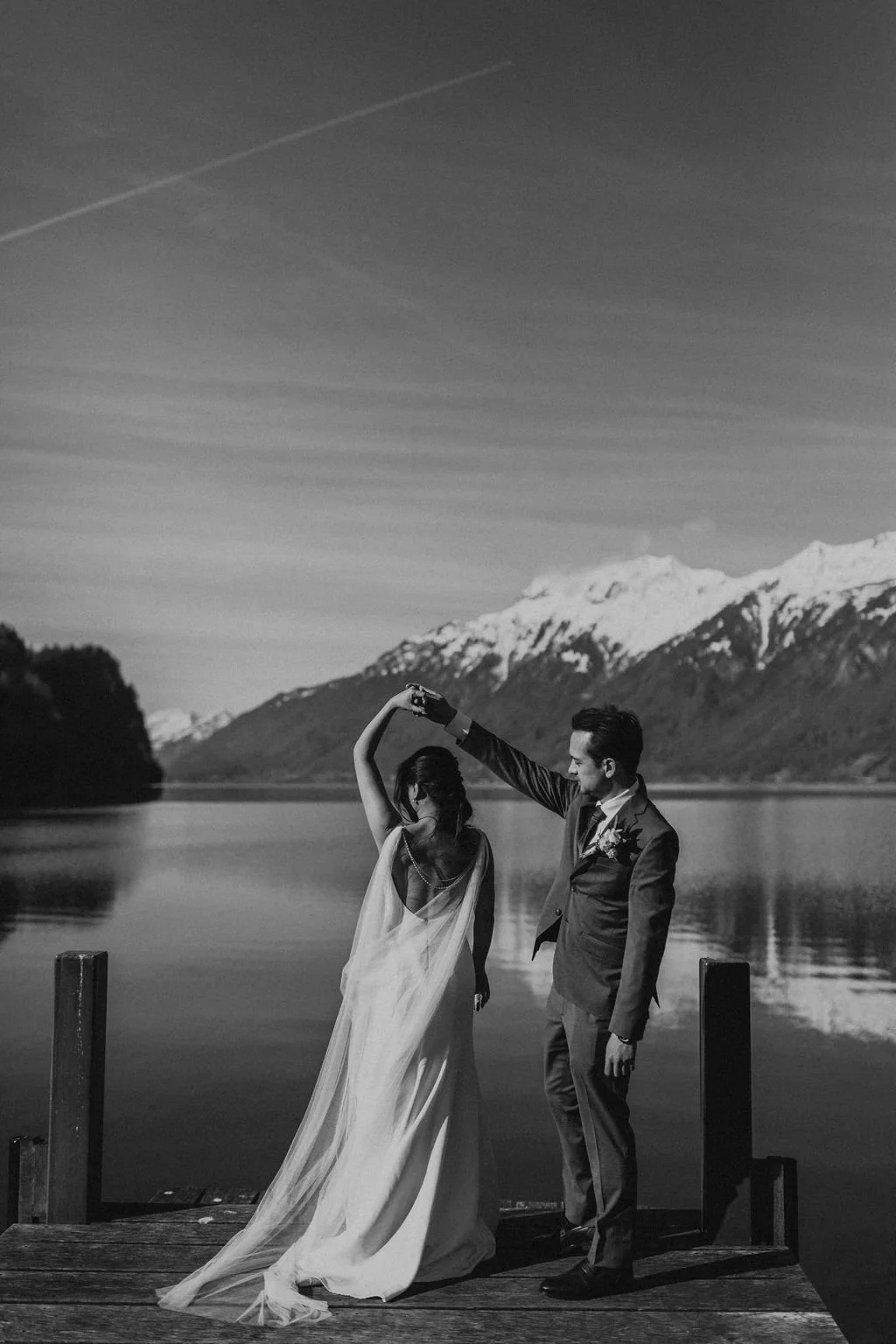 A black-and-white photo of a couple dancing on a dock by a lake with snow-capped mountains in the background, and a clear sky.