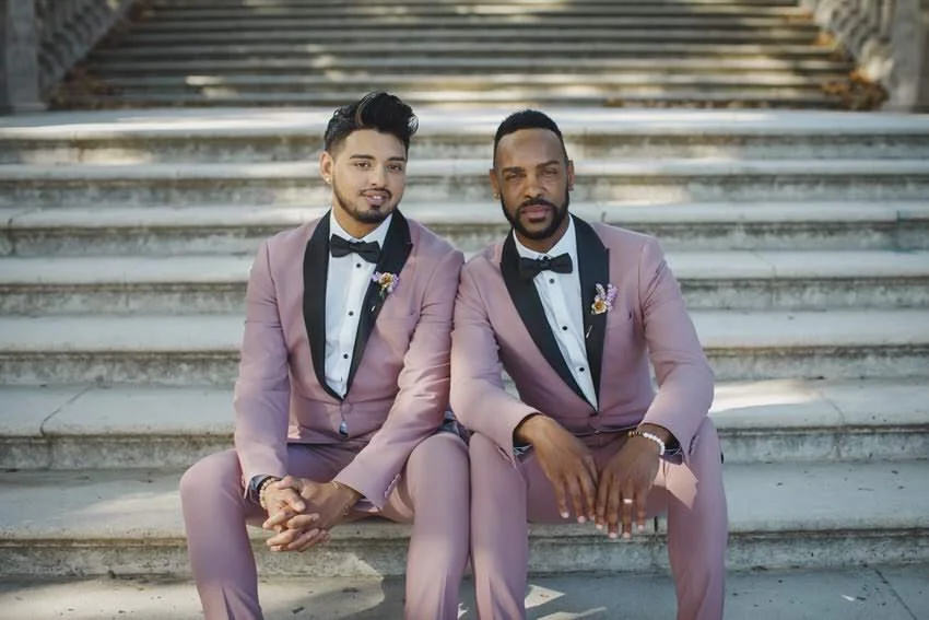 Two men sitting on outdoor stairs, wearing matching pink suits with black lapels and bow ties, posing for a photo.