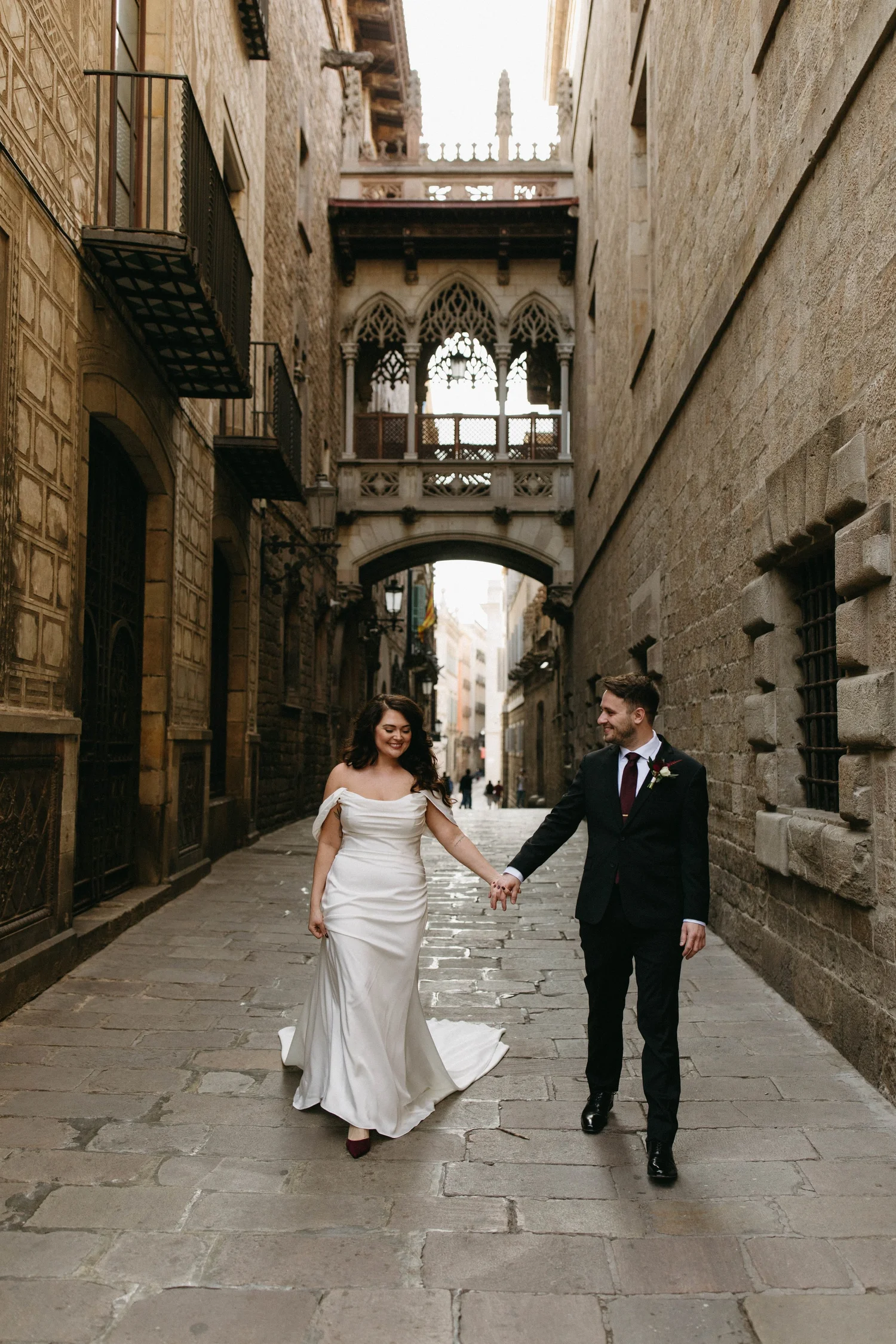A bride and groom walking hand in hand through a historic narrow cobblestone street, smiling at each other, with old stone buildings and ornate architecture in the background.