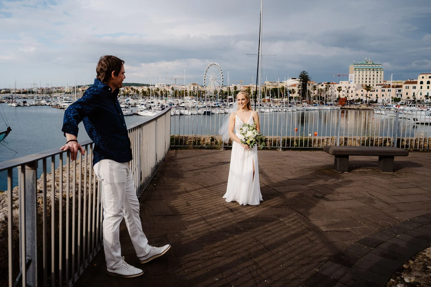 A man and woman standing outdoors near a marina with boats, a ferris wheel, and buildings in the background. The woman is in a wedding dress holding a bouquet, smiling at the man, who is casually dressed and looking at her.