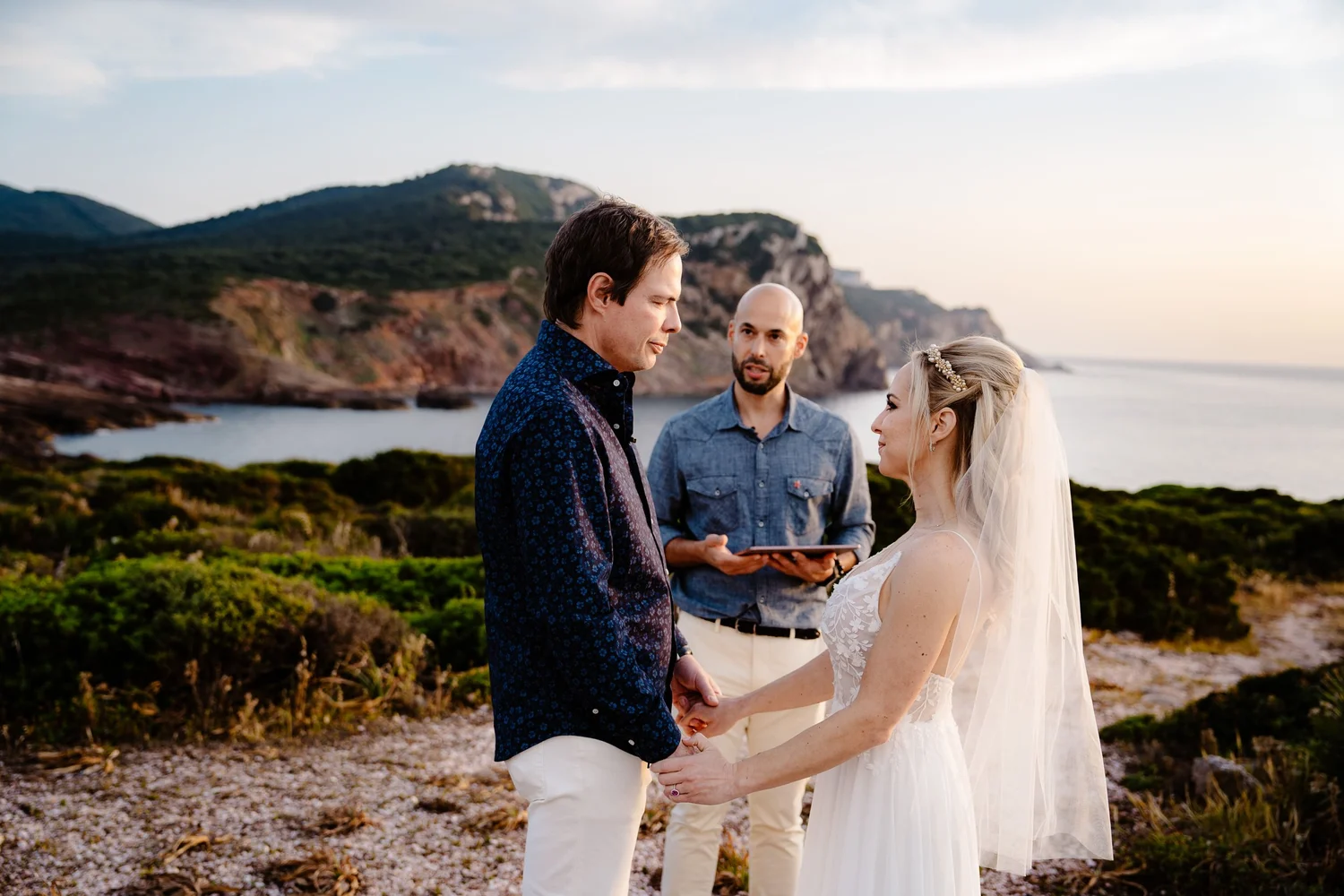 A couple getting married on a rocky beach at sunset, with a man officiating and a scenic view of hills and water in the background.