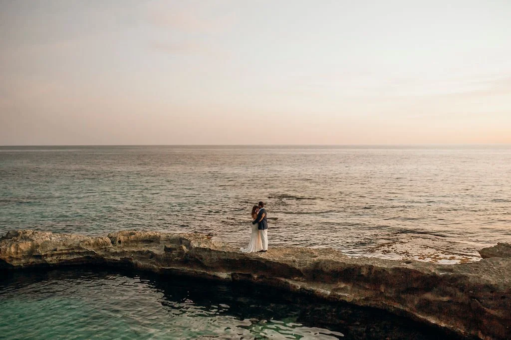 A couple dressed in formal attire standing on rocky terrain by the ocean, embracing with the sea and sunset sky in the background.