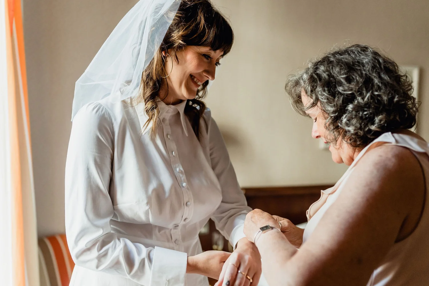 A woman in a white shirt with a veil holding hands with an older woman, sitting close together in a tender moment, likely in a hospital or care setting.