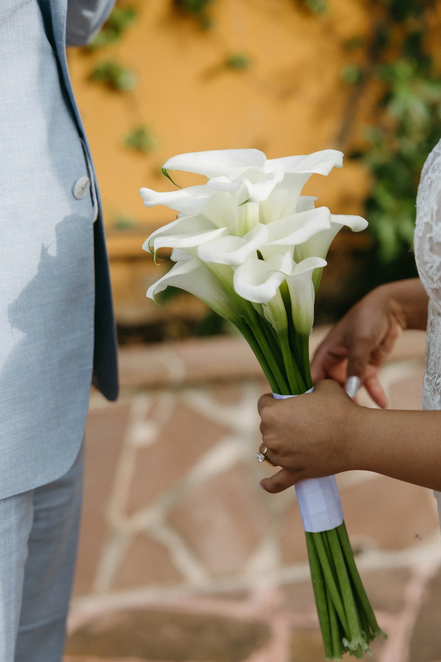 A person holding a bouquet of white calla lilies during a wedding ceremony, with the groom dressed in a light-colored suit visible on the left.