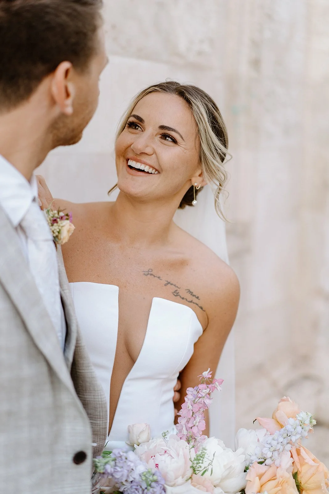 Elopement portrait of a bride with blonde hair and earrings smiling at her groom during a wedding. She has a tattoo on her shoulder that reads "Love you more" and is holding a bouquet of pink and white flowers.
