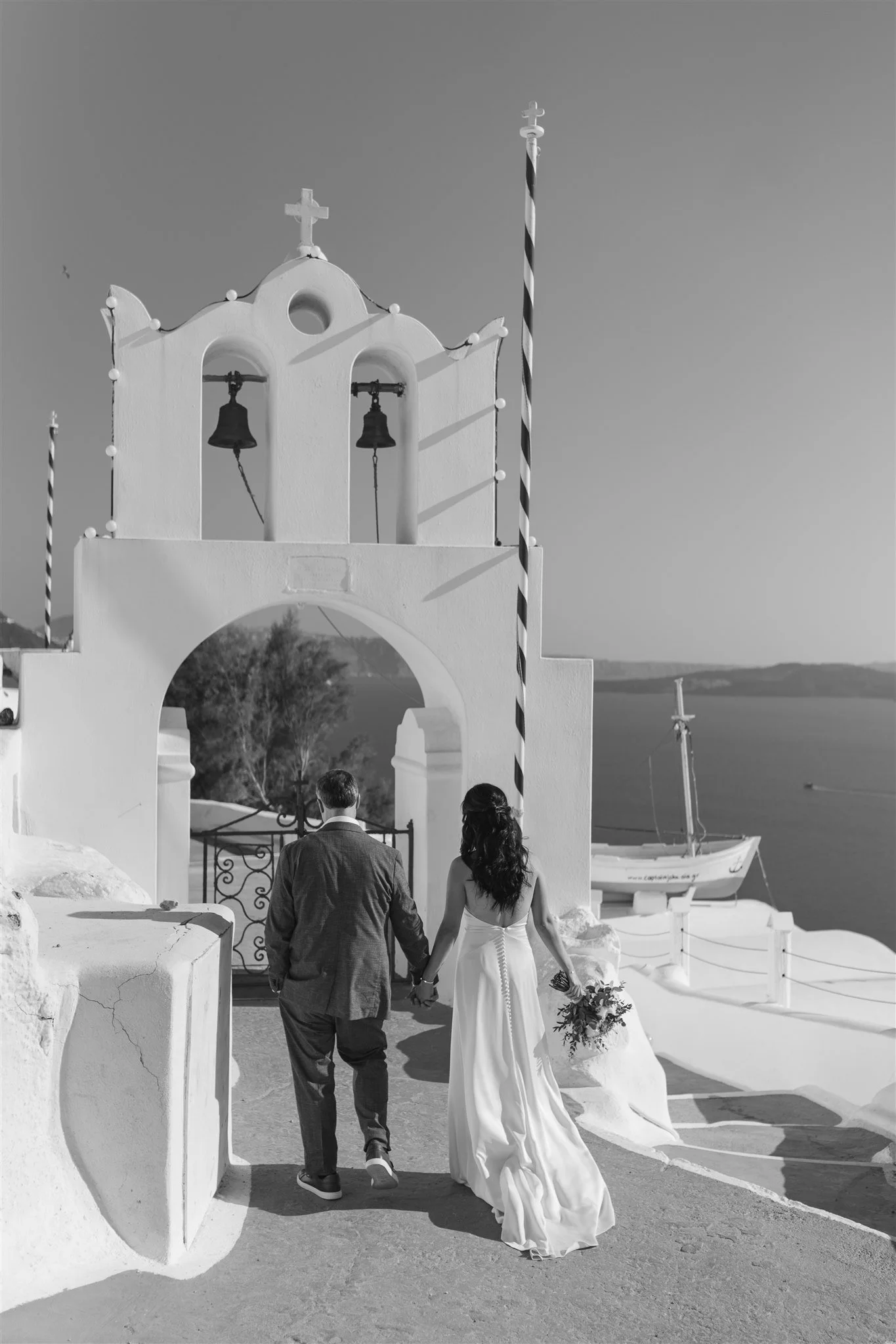 A couple dressed in wedding attire walking hand in hand through a white archway with bells, in a Greek island setting with white buildings and boats in the background.