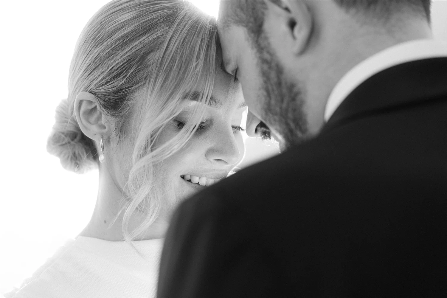 A black and white photo of a couple with their foreheads touching, smiling with eyes closed. The woman has light hair styled in an updo, and the man has short hair and a beard. They are dressed formally.