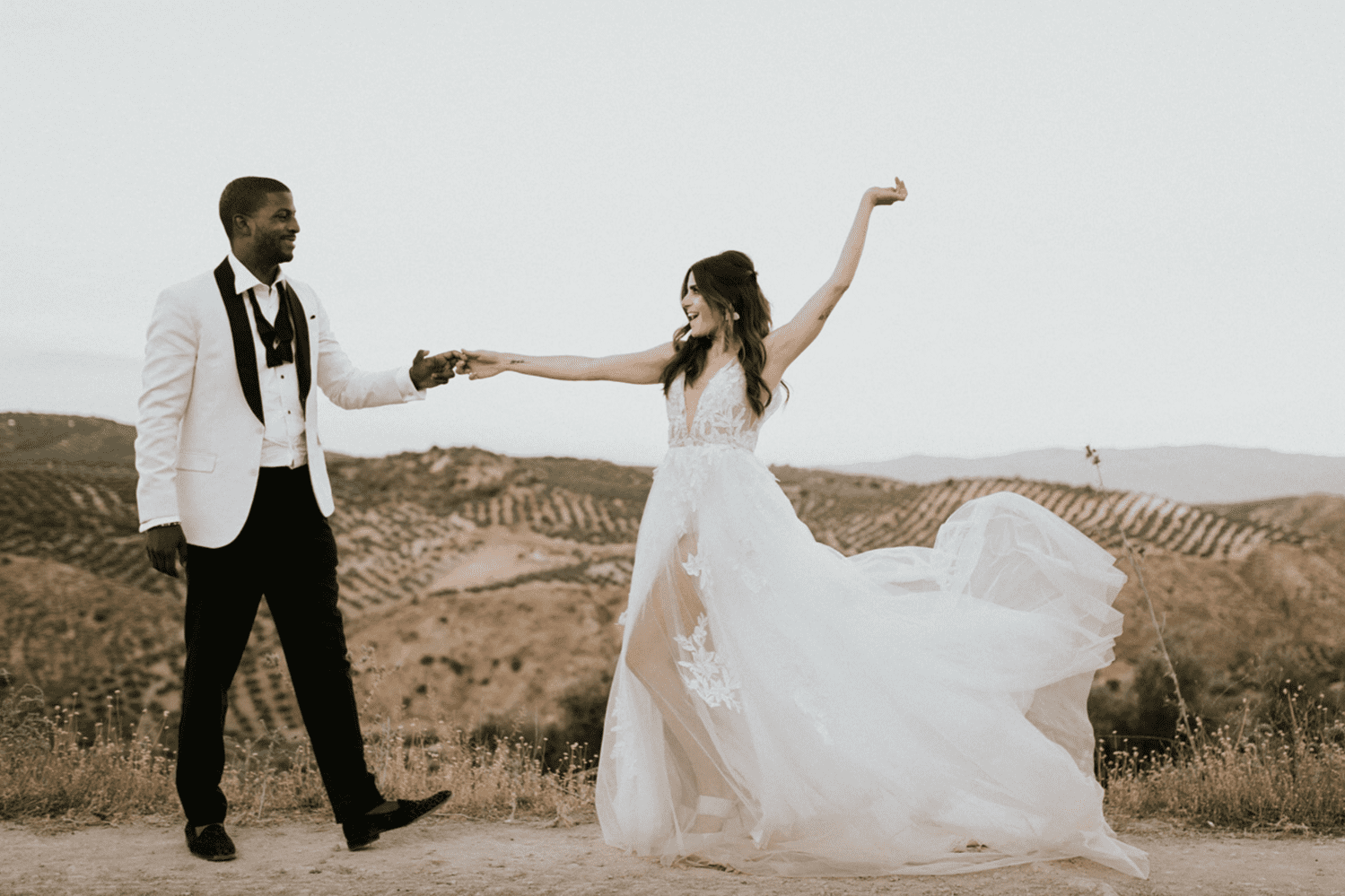 A newlywed couple holding hands and smiling outdoors with hills in the background, the bride is spinning in a white wedding gown.