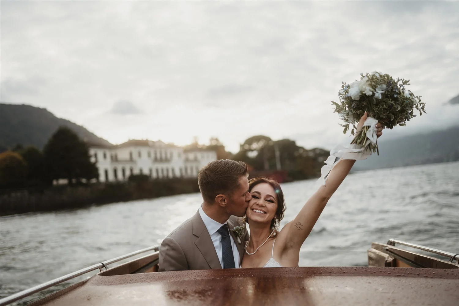 A newlywed couple sharing a kiss on a boat with a person holding a bouquet of flowers. The background shows water, trees, and a large white building.