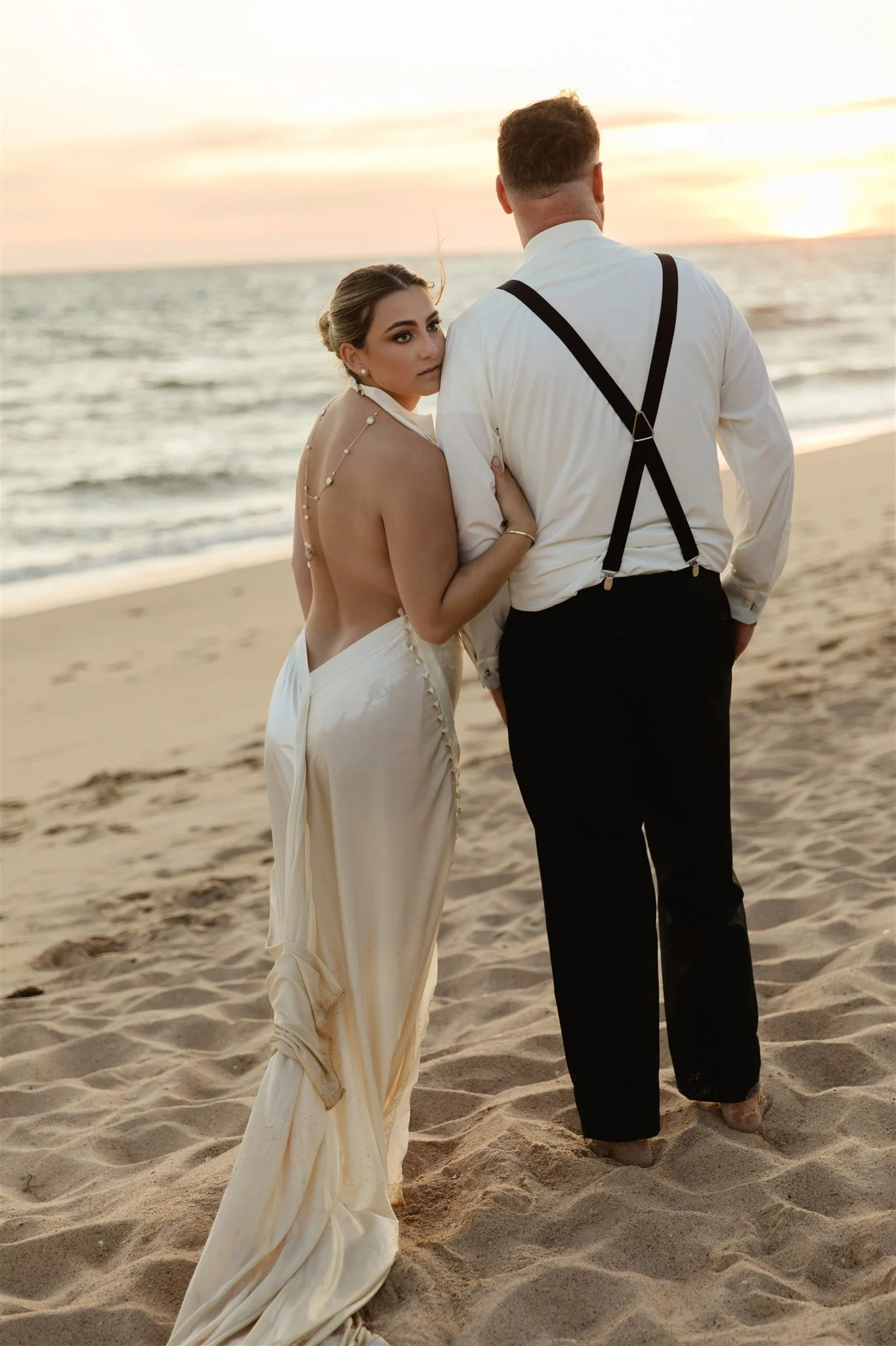 Elopement portrait of a woman in a wedding dress and a man in a white shirt and suspenders standing on the beach during sunset.