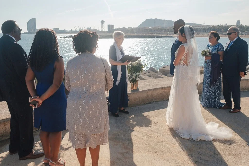 A wedding ceremony taking place outdoors near a body of water with city buildings in the background. A bride in a white lace dress and veil stands alongside her groom in a dark suit, facing an officiant. Guests in formal attire observe the ceremony.