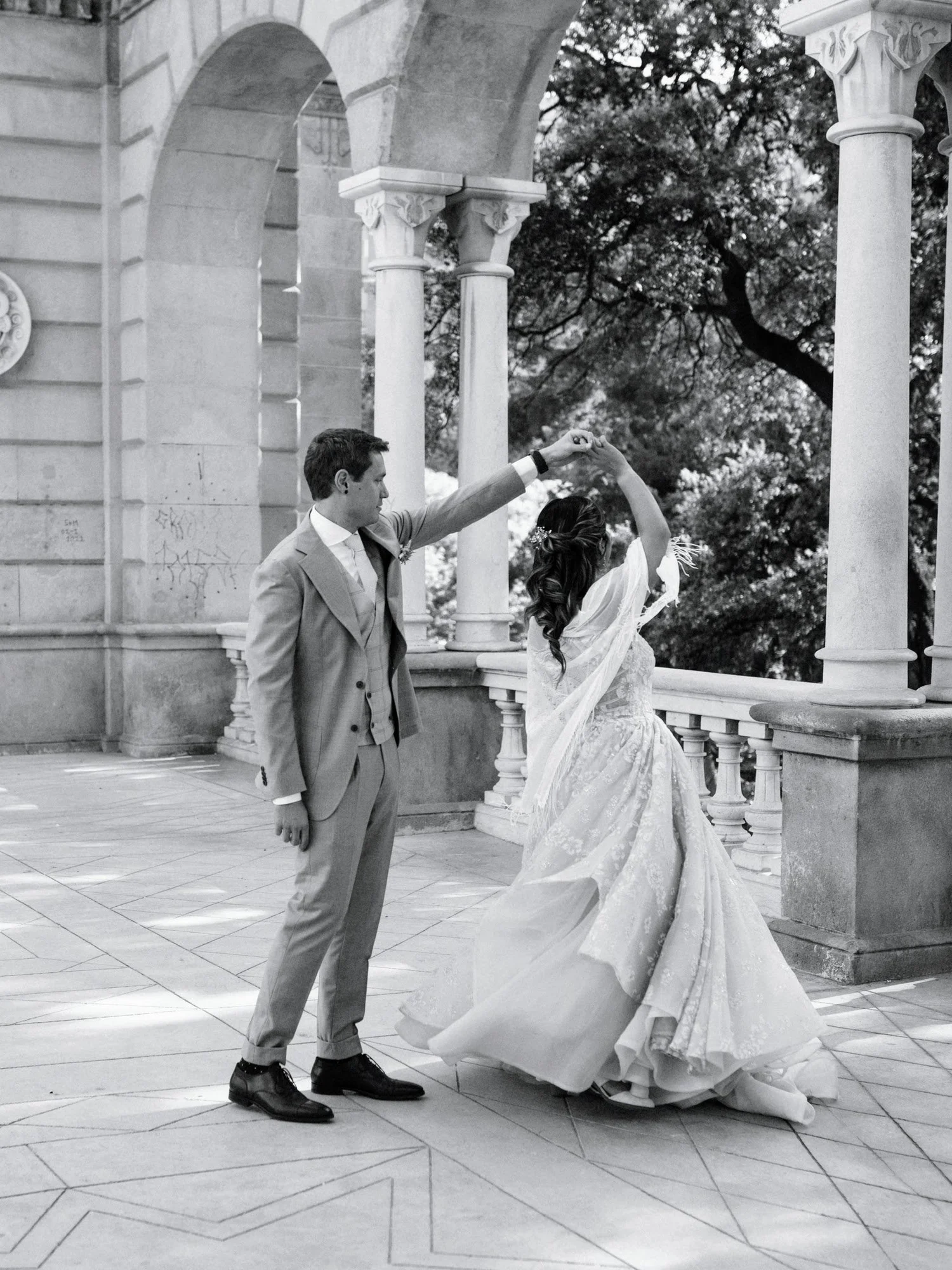 A black-and-white photograph of a bride and groom dancing outside on a balcony with ornate columns and a decorative railing. The groom is dressed in a light suit with dark shoes, and the bride is in a flowing wedding gown with lace detailing, her hair styled with flowers. They are holding hands, with the bride spinning under the groom's arm, surrounded by a scenic background of trees.