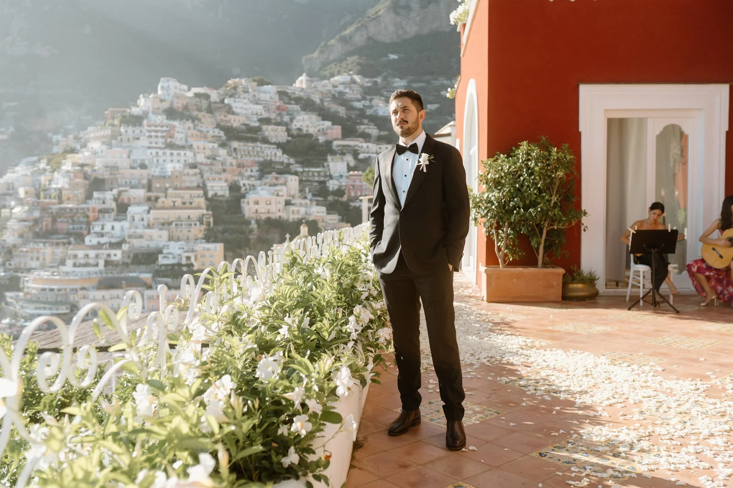 A man in a tuxedo with a white shirt and black bow tie standing on a terrace with a view of a hillside town, white flowers in the foreground, and a warm-colored building.