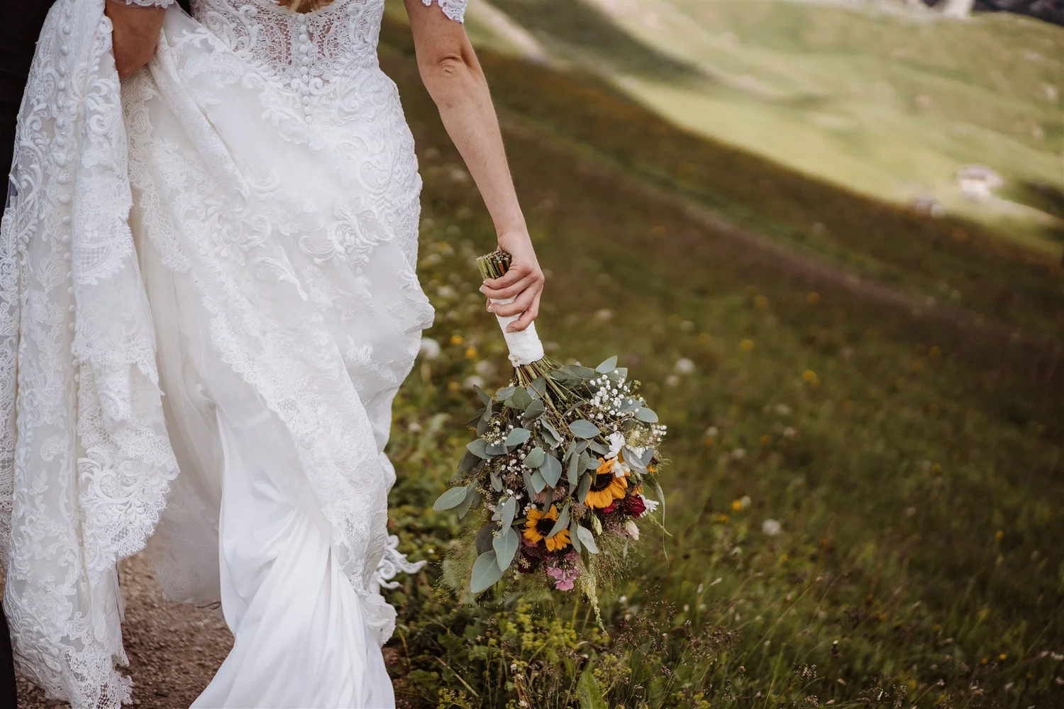 A bride wearing a white lace wedding dress holding a bouquet of flowers walking along a grassy path outdoors.
