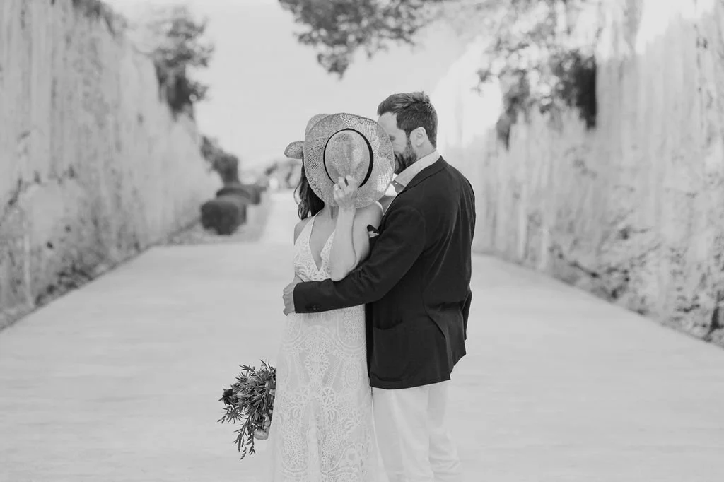A couple dressed in wedding attire sharing a kiss outdoors, with the woman holding a bouquet and wearing a wide-brimmed hat, and the man in a suit.