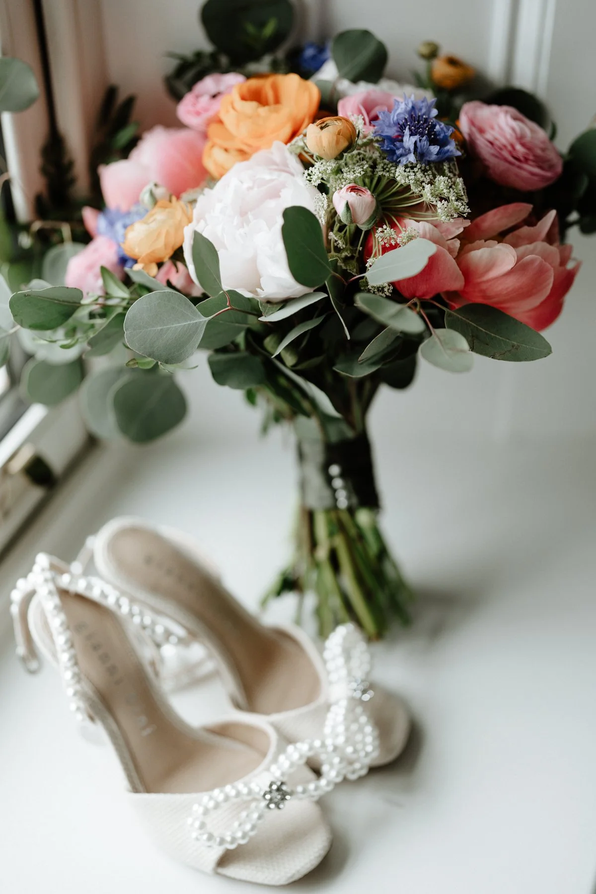 A bouquet of mixed flowers including peonies, ranunculus, and blue cornflowers with eucalyptus leaves, placed near a pair of ivory high-heeled shoes decorated with pearls and rhinestones.