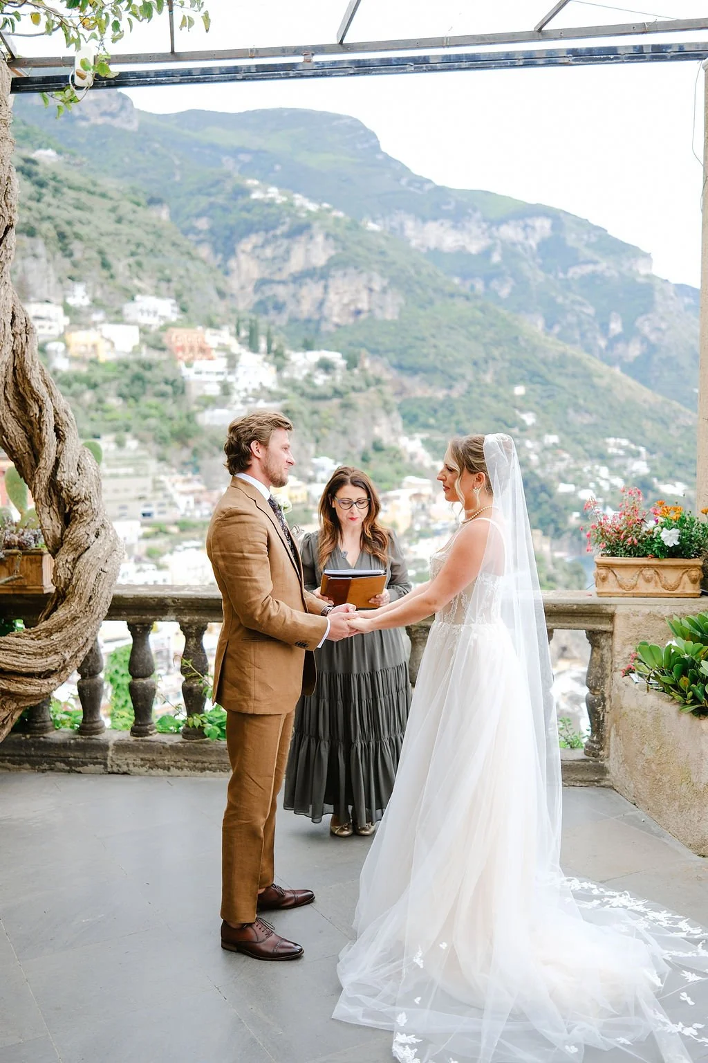 A couple getting married outdoors on a terrace with a mountainous background, the bride in a white wedding dress and veil, the groom in a brown suit, with an officiant standing between them.