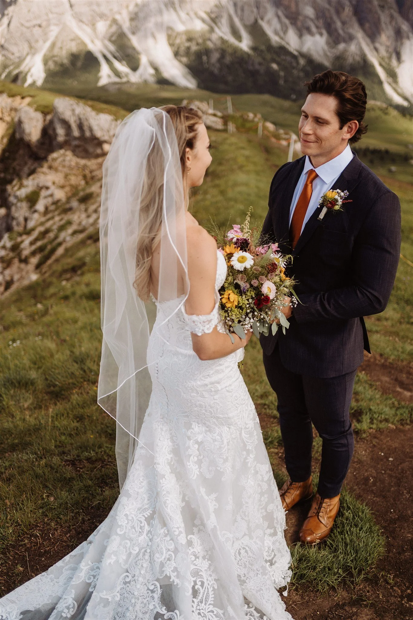 A bride and groom holding hands and smiling at each other outdoors with green grass and mountainous terrain in the background.