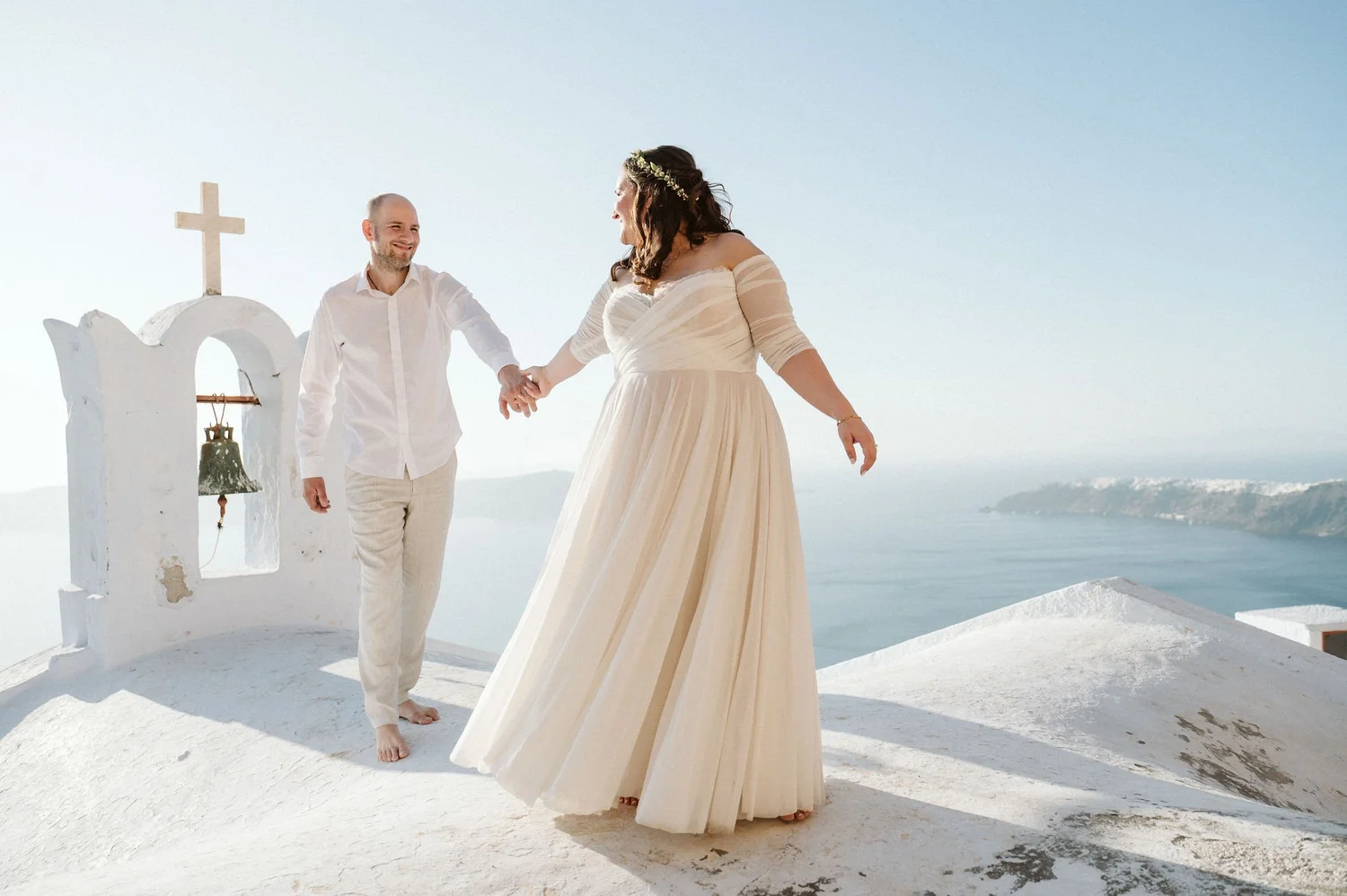 A newlywed couple holding hands and smiling during their wedding ceremony on a white hillside overlooking the ocean in Santorini, Greece. The bride is wearing a long, flowing cream-colored gown with off-the-shoulder sleeves and a floral crown, while
