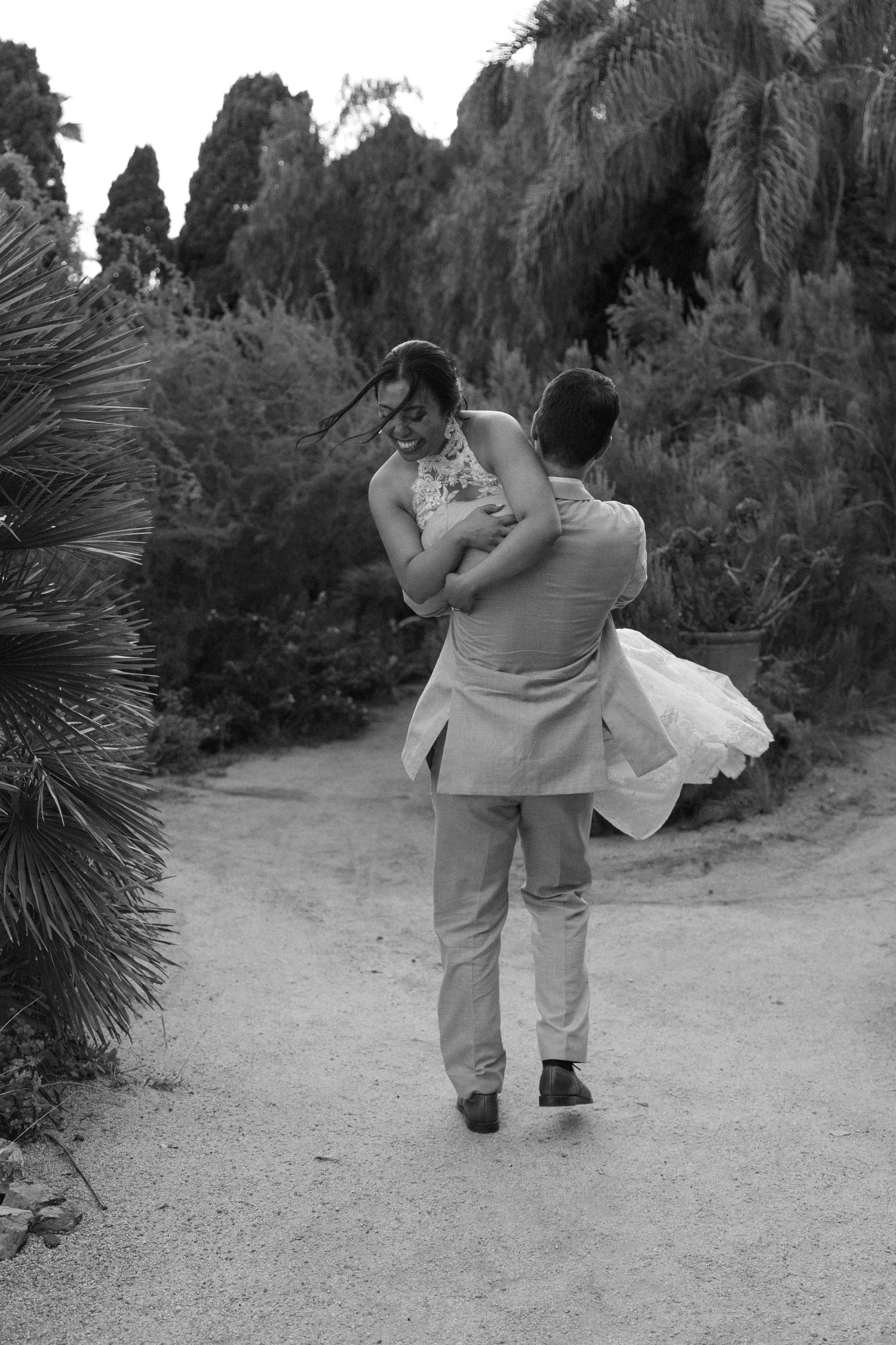 A couple, with the woman in a dress and the man in a suit, is dancing together outdoors. The woman is smiling and the man is holding her. They are walking along a dirt path surrounded by trees and plants.