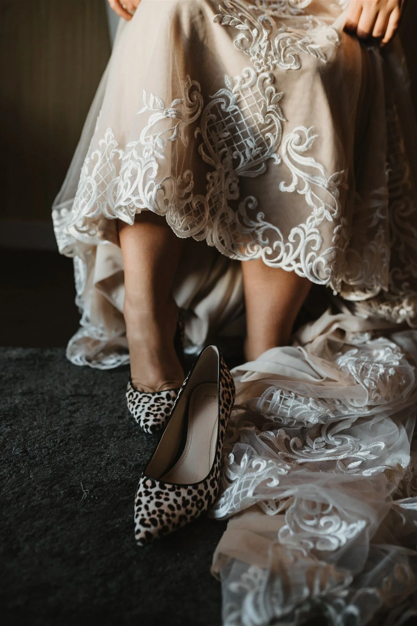 Close-up of a woman putting on a leopard print high heel shoe in front of a wedding dress with intricate embroidery and lace.