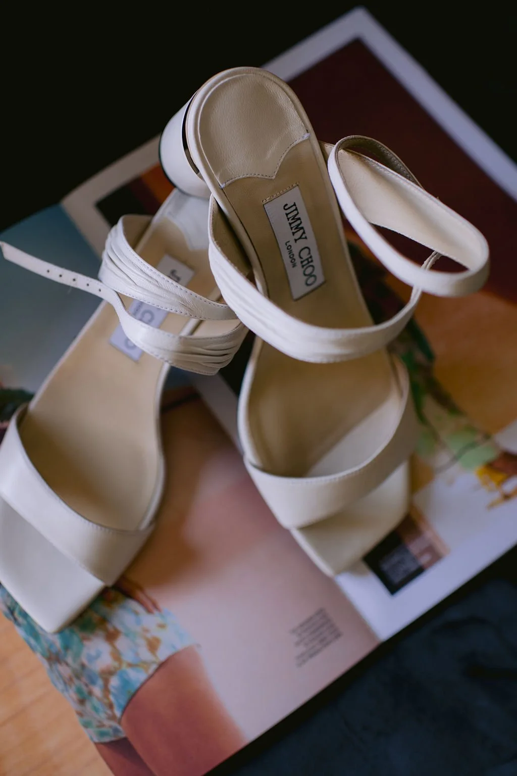 A pair of white high-heeled sandals with ankle straps placed on top of a magazine on a wooden surface.