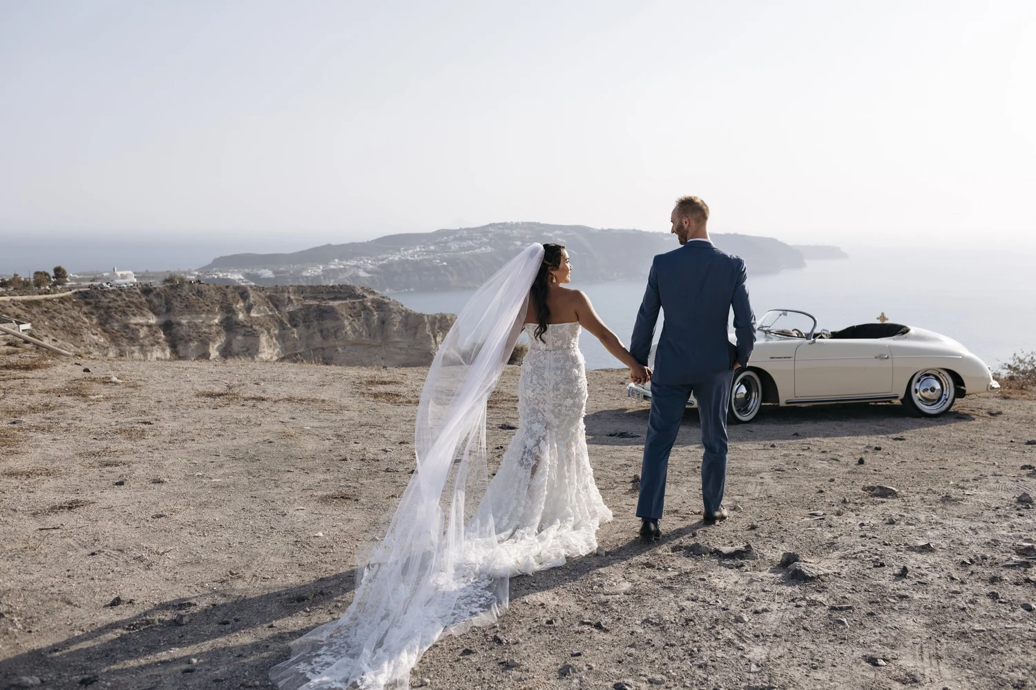 A bride and groom holding hands in a scenic outdoor setting with a vintage car and ocean view in the background.