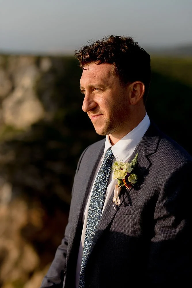 A man dressed in a gray suit with a white shirt and patterned tie, wearing a boutonniere, standing outdoors in sunlight with a blurred natural background.