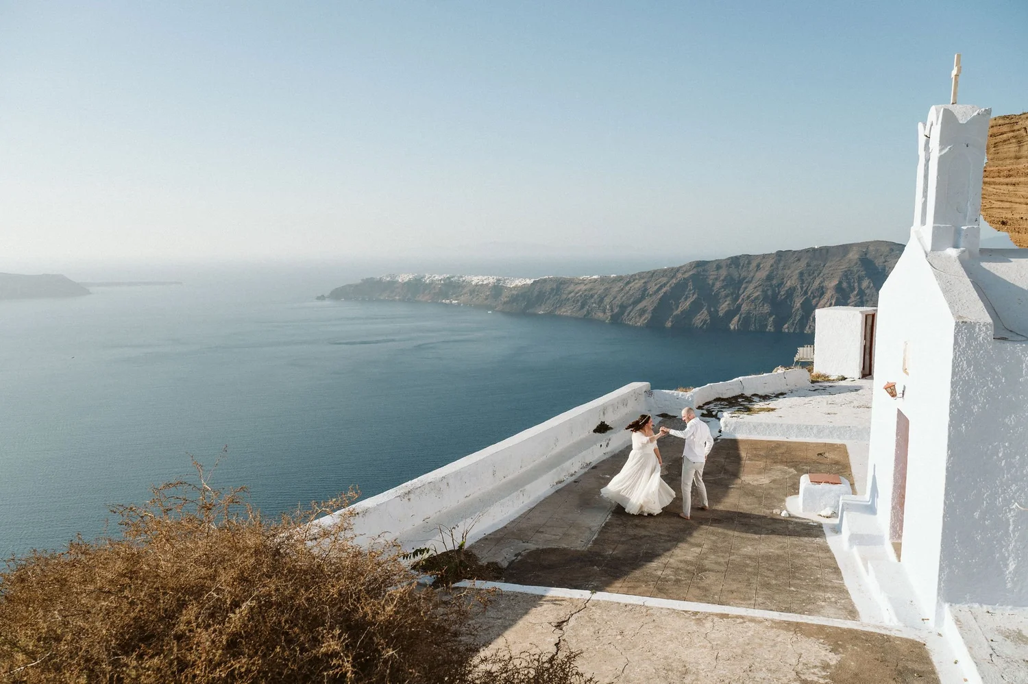 A couple dancing on a terrace overlooking the sea with white buildings and cliffs in the background, during a sunny day.