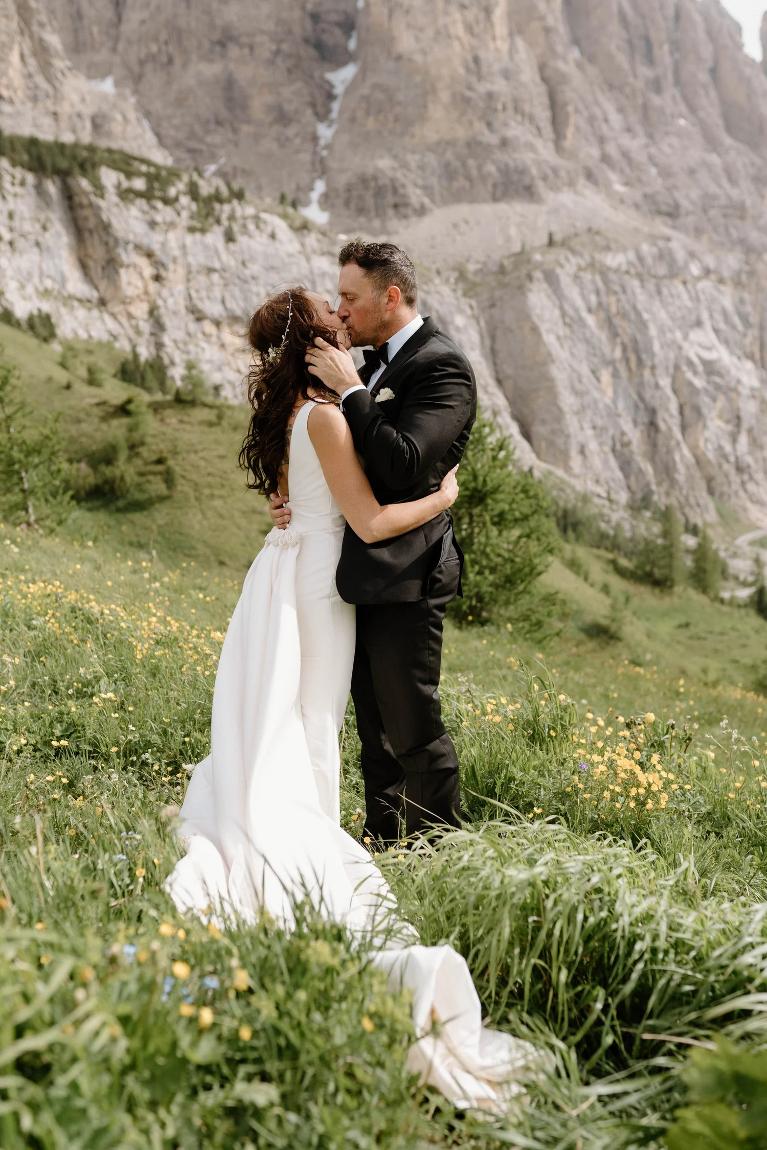 A bride and groom sharing a kiss outdoors on a grassy field with wildflowers, mountains in the background, during a wedding.
