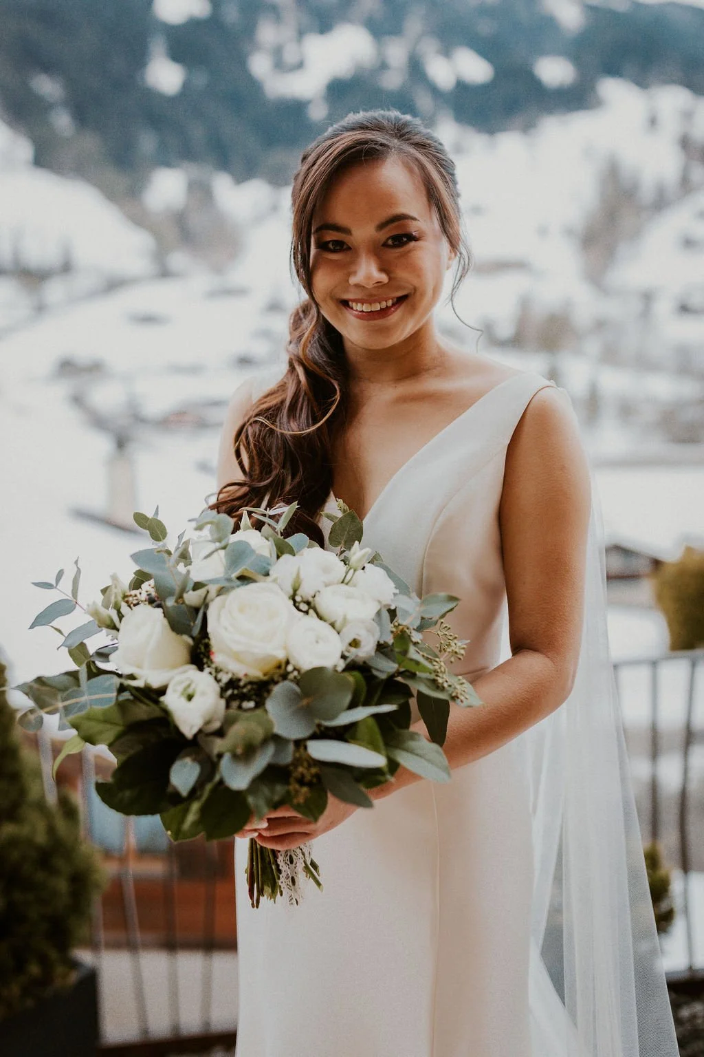 A smiling bride with long, wavy brown hair holding a bouquet of white roses and greenery, standing outdoors with a snowy hillside and lake in the background.