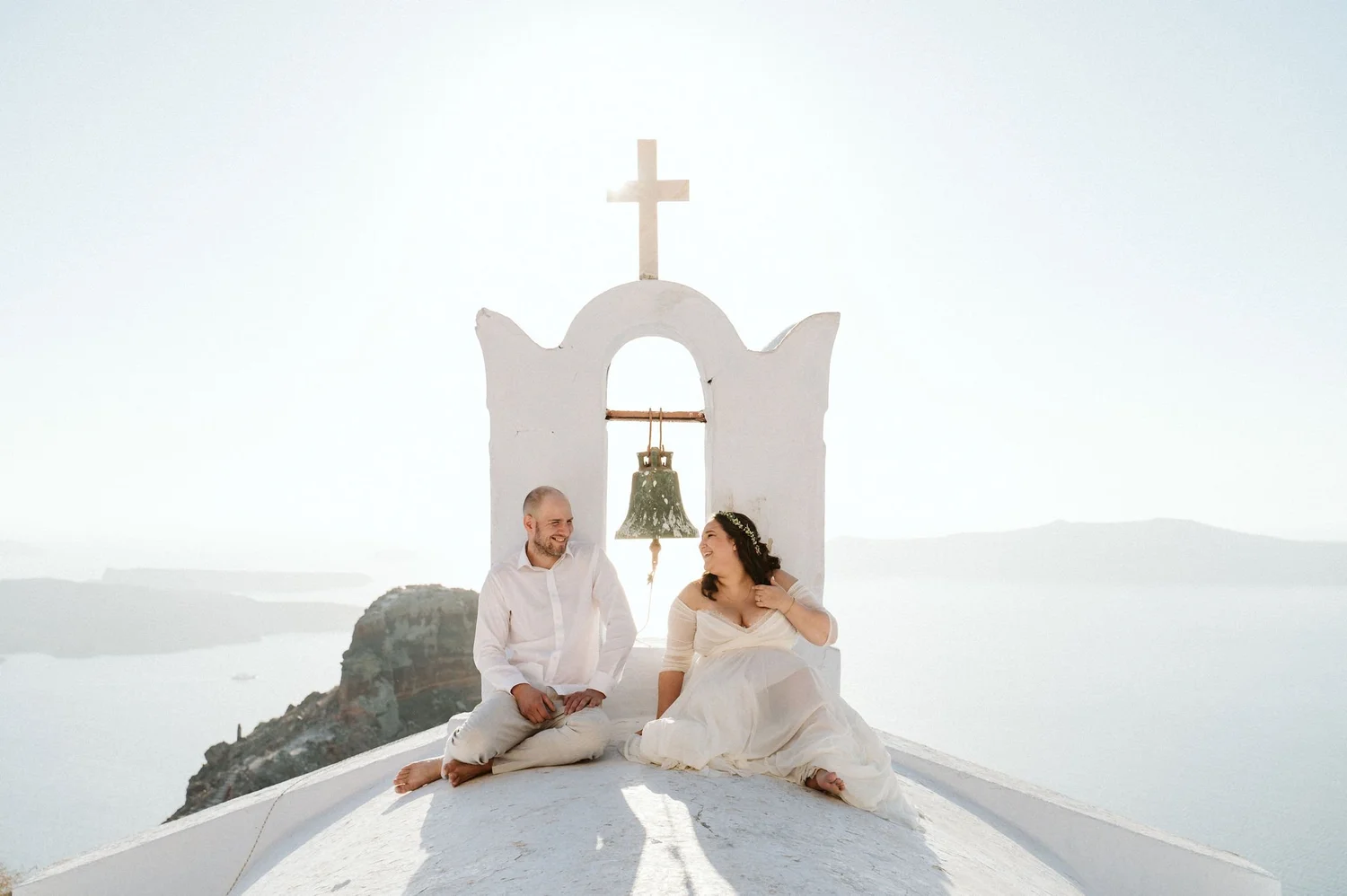 A couple dressed in white, sitting on the roof of a white building with a bell and a cross, overlooking a calm, sunny landscape with mountains in the distance.