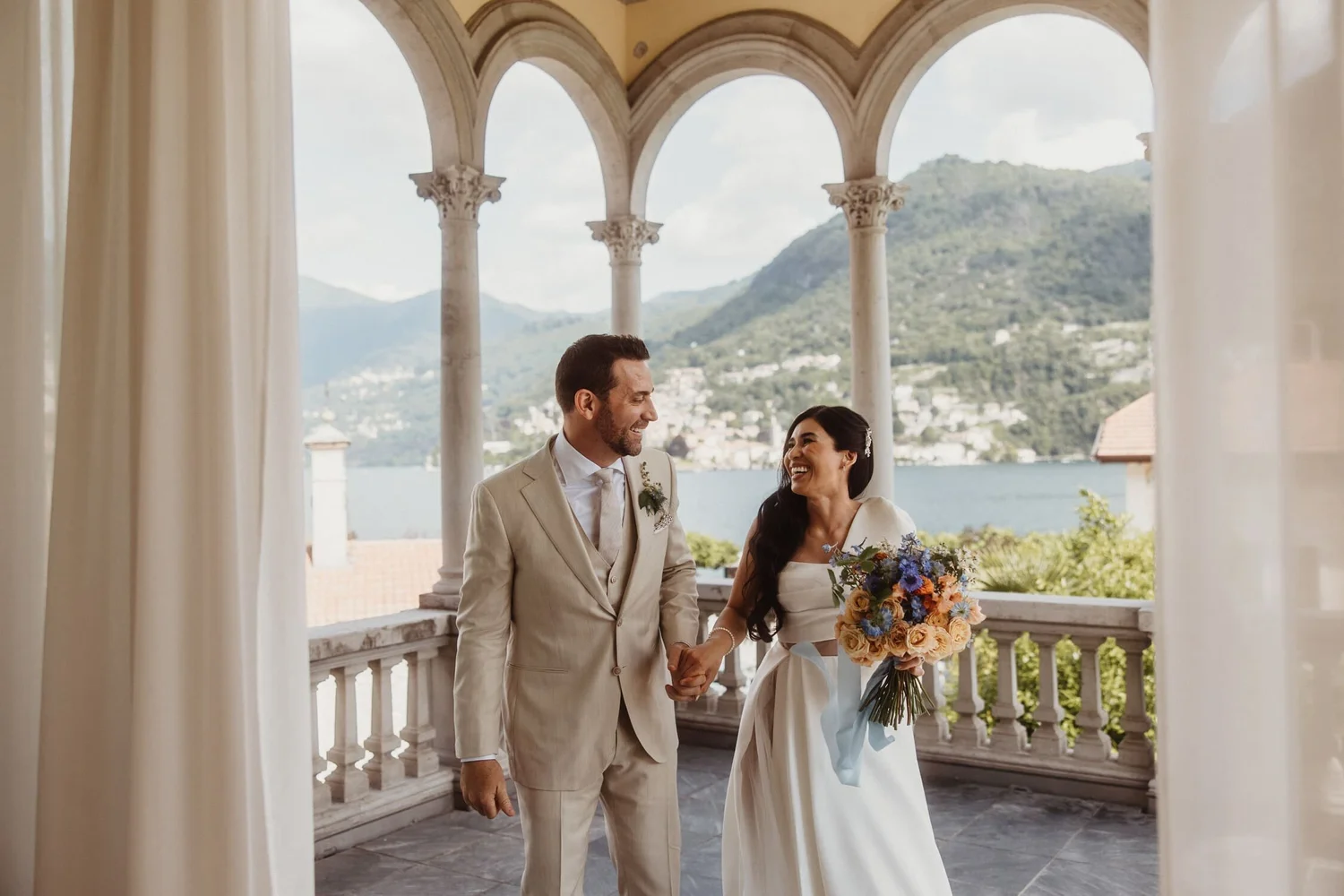 A newlywed couple holding hands and smiling at each other on a balcony with scenic mountain views in the background. The man is dressed in a beige suit and the woman in a white wedding gown holding a bouquet of flowers.