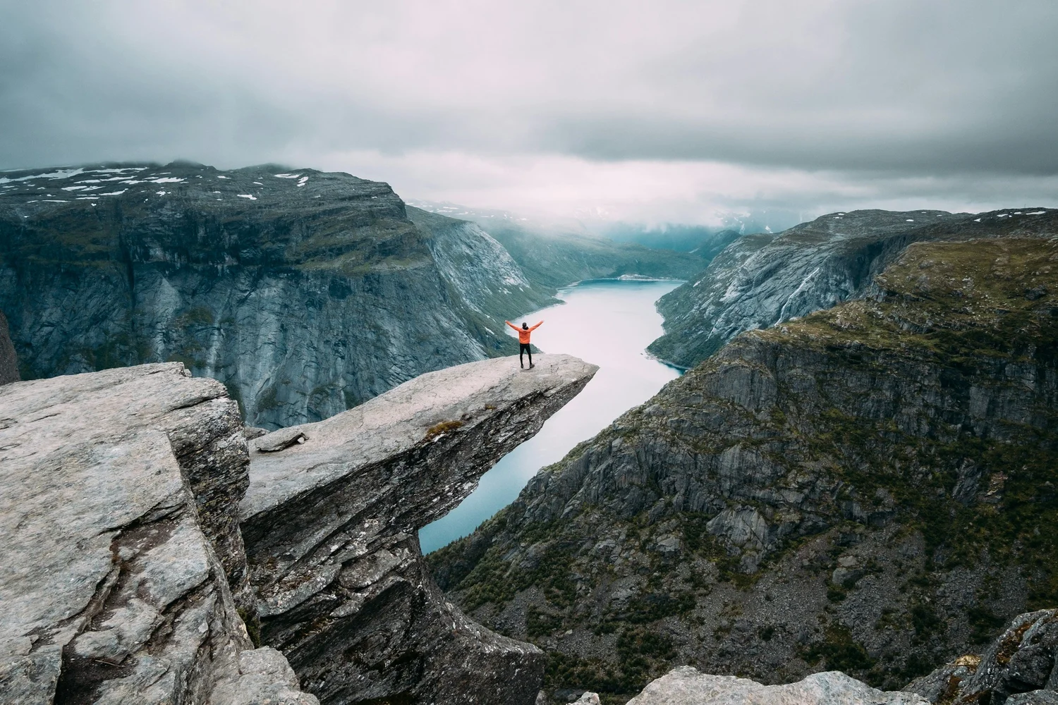 Person standing on a large rock ledge overlooking a deep river gorge with cliffs on both sides and snow patches on the mountains in the distance, under a cloudy sky.