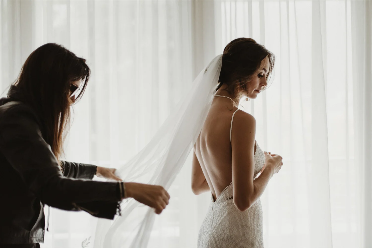 A bride getting ready for her wedding, assisted by a stylist, with a sheer white curtain in the background.