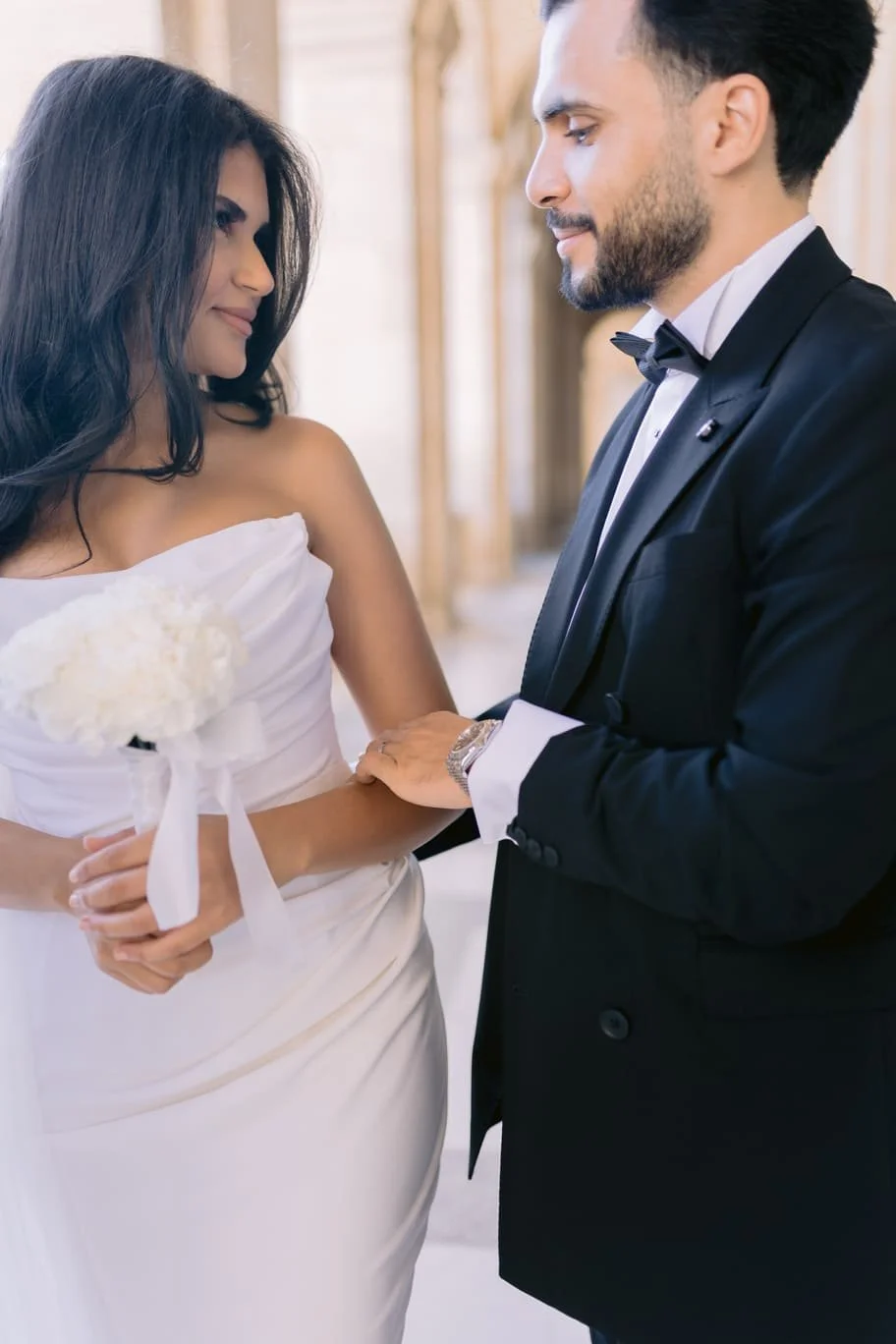A bride and groom sharing a tender moment during their wedding ceremony, with the bride holding a white bouquet and wearing a strapless white wedding dress, and the groom dressed in a black tuxedo with a bow tie.