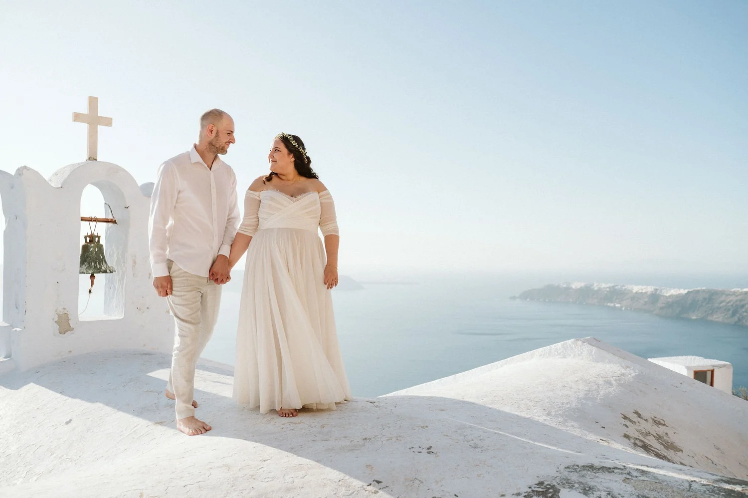 A couple dressed in wedding attire holding hands and looking at each other on a white cliff overlooking the ocean in Santorini, Greece, with a church bell and cross nearby.