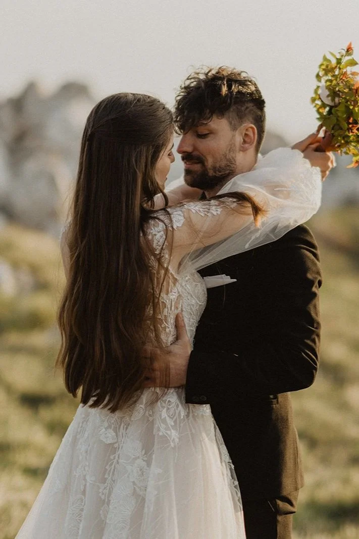 A wedding couple stands closely together outdoors, with the woman embracing the man. The woman wears a white lace wedding dress, and the man wears a dark suit. The woman holds a bouquet of flowers behind his head, and they gaze at each other lovingly.