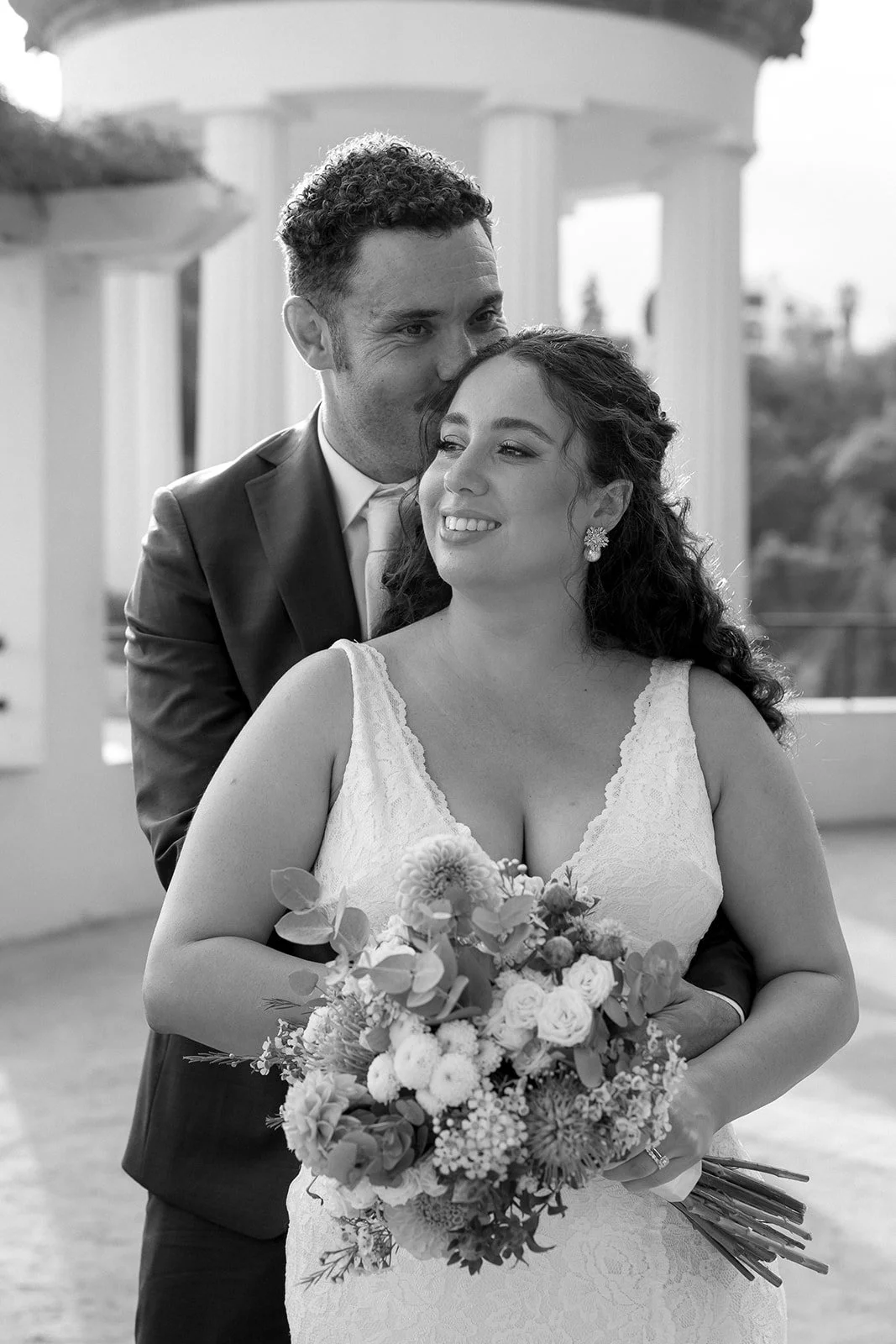 A black and white photo of a bride holding a bouquet of flowers, with a groom standing behind her, kissing her forehead. They are outdoors with classical pillars in the background.