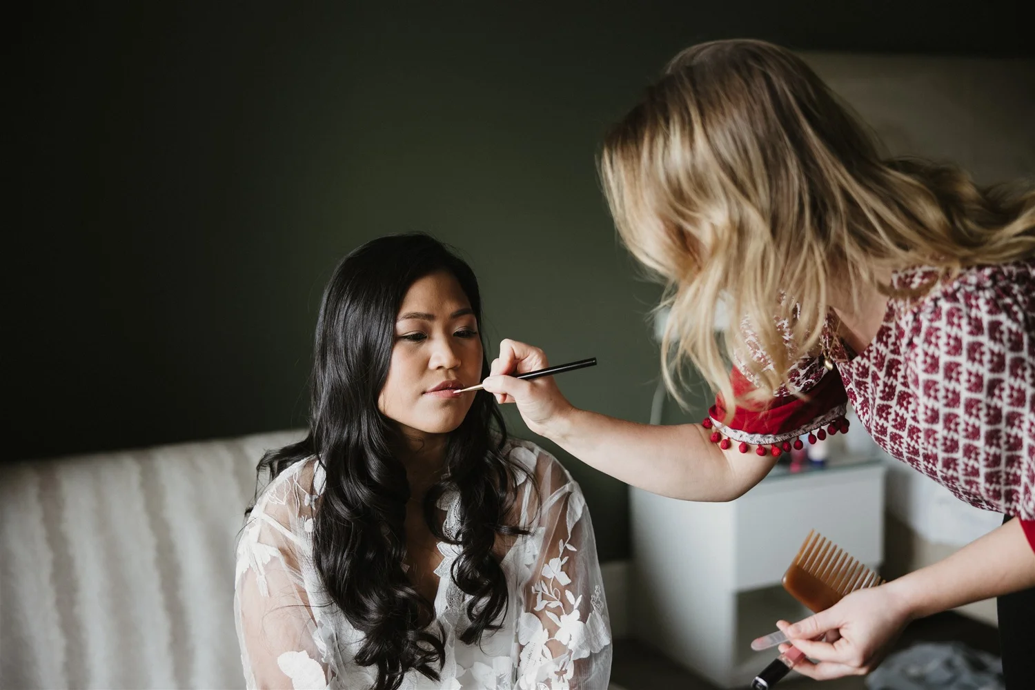 A woman with long black hair and a white floral blouse gets her makeup done by a professional makeup artist with blonde hair and a red and white patterned top.