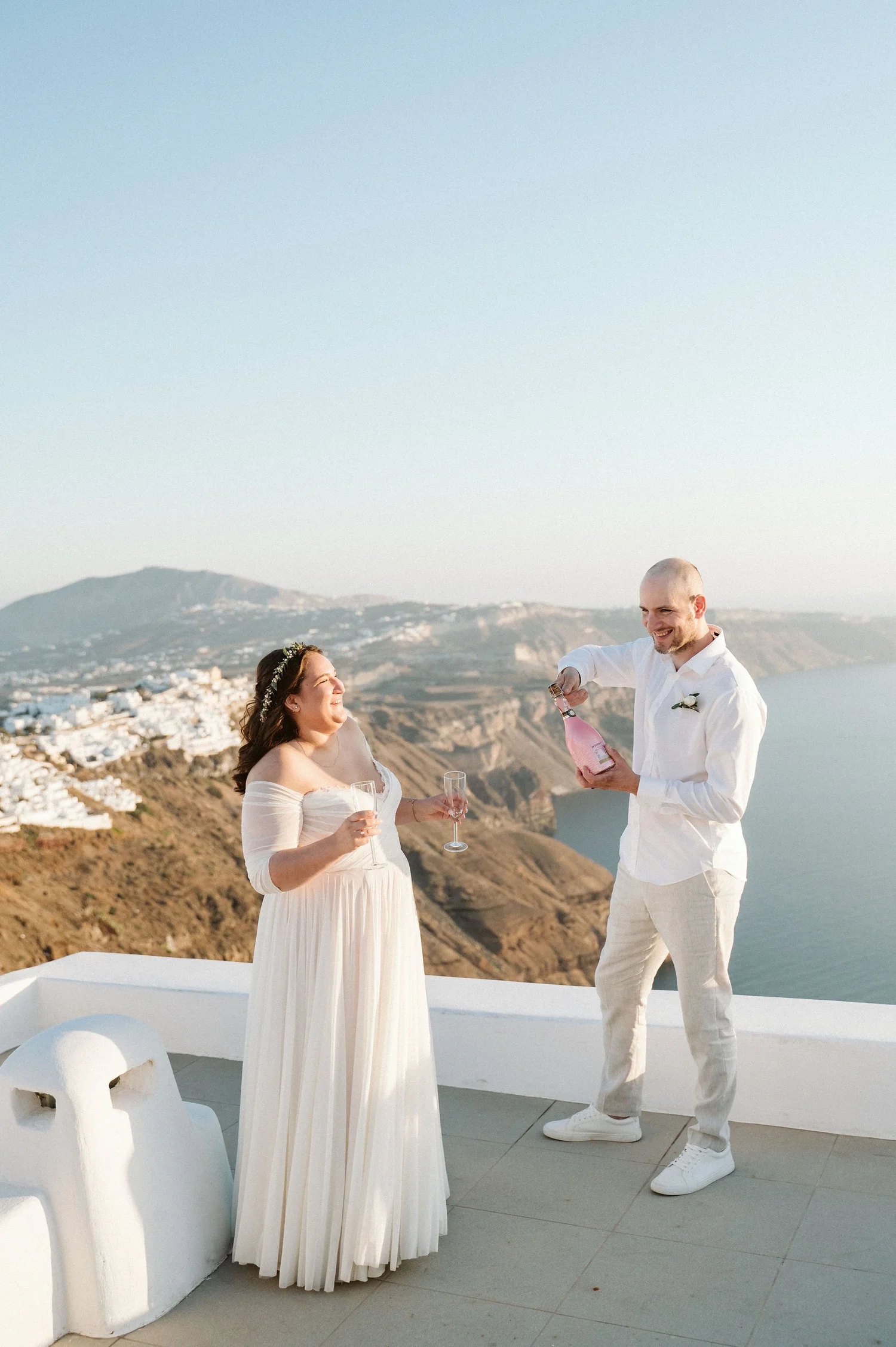A bride and groom celebrating on a terrace overlooking a coastal landscape with white buildings and hills. The bride is wearing a white off-shoulder dress and a floral headband, holding a glass of champagne. The groom is in white pants and shirt, ope